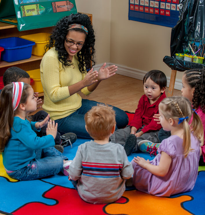 An early childhood educator leads a circle activity with young children on a colourful classroom rug.